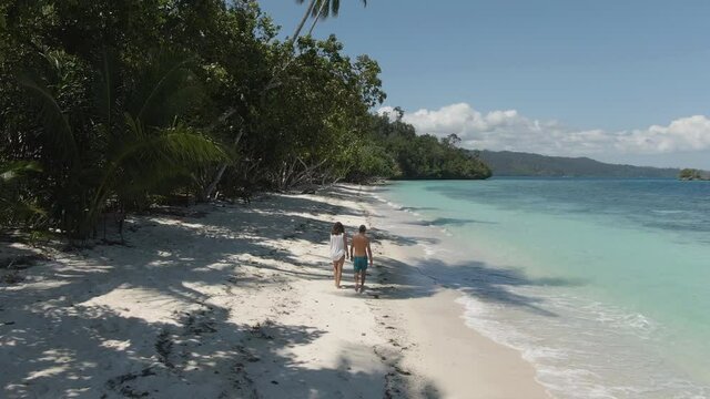 Aerial 4K view following a couple walking on a stunning paradise beach in Gam island, Raja Ampat, Papua, Indonesia