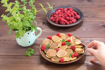 The hand with the fork reaches for the Pancake cereals. Red ripe fresh raspberries in a brown plate and a bunch of Melissa in a mug on a dark wooden table. Delicious Breakfast