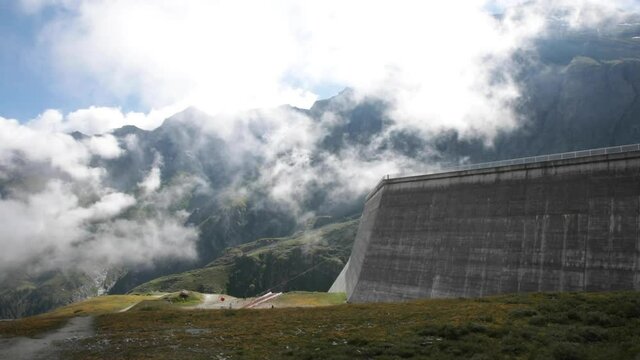 Large dam in Switzerland in the middle of the mountain with some rolling clouds over green fields with the sun in frame. 50fps