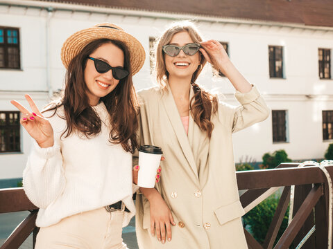 Two young beautiful smiling hipster girls in trendy white sweater and coat. Sexy carefree women posing on street background in hat and sunglasses. Positive models having fun and drinking coffee