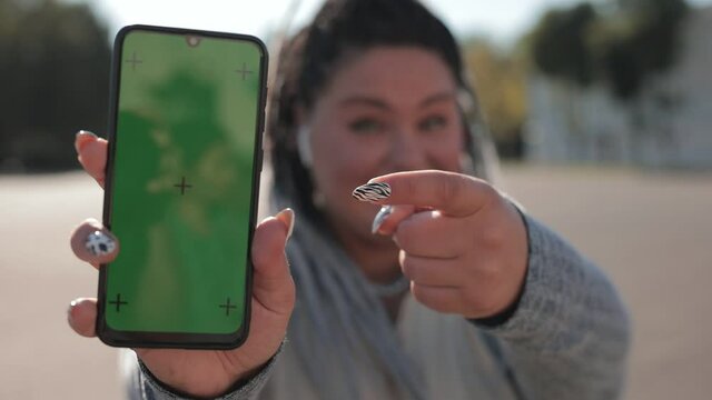 Plump cheerful woman with dreadlocks points at a phone with a green screen with motion capture points. Concept of advertising an app for weight loss, food, sports and running