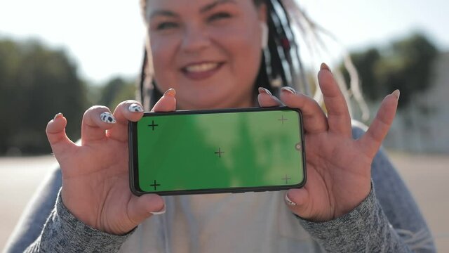 Plump cheerful woman with dreadlocks points at a phone with a green screen with motion capture points. Concept of advertising an app for weight loss, food, sports and running