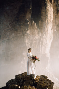 Happy bride with flower bouquet standing on stone cliff and enjoy the sunset in mountain
