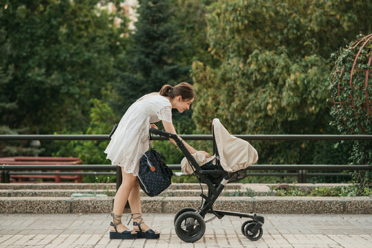 Mother in white dress walking with baby carriage in park.