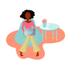 African woman working on her laptop sitting on a beanbag chair in front of the coffee table with flowers.