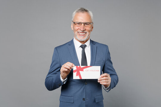Successful elderly gray-haired mustache bearded business man in classic blue suit shirt tie isolated on grey wall background studio. Achievement career wealth business concept. Hold gift certificate.