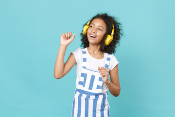 Funny little african american kid girl 12-13 years old in striped clothes isolated on blue background in studio. Childhood lifestyle concept. Mock up copy space. Listen music with headphones dancing.