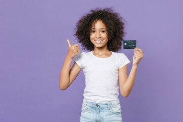 Smiling little african american kid girl 12-13 years old in white t-shirt isolated on violet wall background. Childhood lifestyle concept. Mock up copy space. Hold credit bank card, showing thumb up.