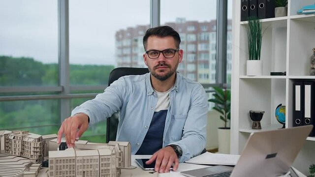 Handsome confident creative young bearded architect in glasses working in design office with wooden model of new buildings and looking at camera with lucky smile