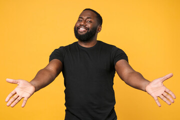 Smiling african american man guy football fan in casual black t-shirt isolated on yellow background studio. People lifestyle concept. Mock up copy space. Standing with outstretched hands for hugs.