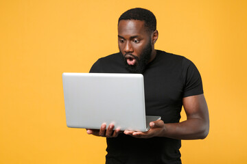 Shocked african american man guy in casual black t-shirt isolated on yellow background studio portrait. People sincere emotions lifestyle concept. Mock up copy space. Working on laptop pc computer.
