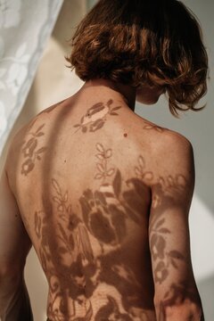 Androgynous man standing shirtless beside window with lace curtain in daylight