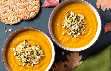 Delicious pumpkin soup with seeds in bowls and dry autumn leaves on a dark table top view. Autumn food. Thanksgiving food.