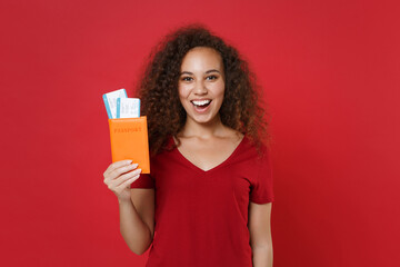 Cheerful young african american woman girl in casual t-shirt isolated on red background. Passenger traveling abroad to travel on weekends getaway. Air flight journey concept. Hold passport tickets.