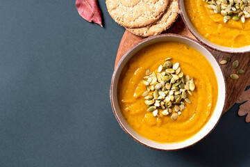 Pumpkin soup with pumpkin seeds in bowls and dry autumn leaves on a dark gray background top view. Free space for text. Copy space. Flat lay. Autumn food. American cuisine.