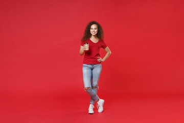 Full length portrait of smiling young african american girl in casual t-shirt isolated on red background studio portrait. People lifestyle concept. Mock up copy space. Hold paper cup of coffee or tea.