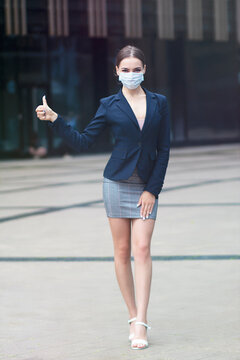 Vertical photo, portrait of beautiful business woman, young gorgeous girl in medical protective mask on face standing, show thumb up, like gesture. Coronavirus, virus, epidemic, covid-19 concept