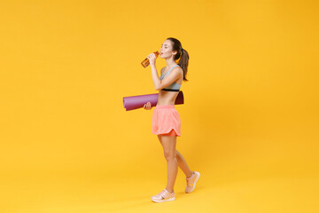 Full length portrait of beautiful young fitness woman girl in sportswear working out isolated on yellow background studio. Workout sport motivation lifestyle concept. Hold yoga mat, bottle of water.