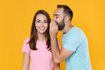 Naklejka premium Smiling young couple friends guy girl in blue pink t-shirts isolated on yellow background studio portrait. People lifestyle concept. Mock up copy space. Whispering secret behind hand, sharing news.