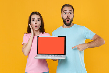 Shocked couple friends guy girl in blue pink t-shirts isolated on yellow background. People lifestyle concept. Mock up copy space. Pointing index finger on laptop pc computer with blank empty screen.