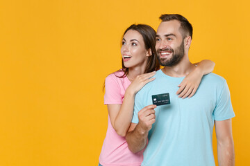 Smiling young couple two friends guy girl in blue pink t-shirts posing isolated on yellow wall background. People lifestyle concept. Mock up copy space. Hold credit bank card, looking aside, hugging.