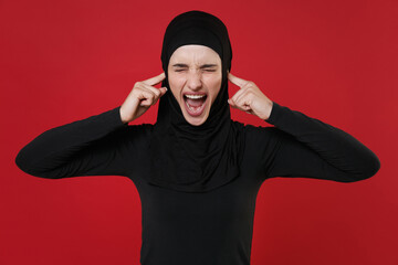 Crazy young arabian muslim woman in hijab black clothes isolated on red background. People religious lifestyle concept. Mock up copy space. Covering ears with fingers, keeping eyes closed, screaming.