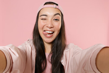 Close up of blinking asian woman girl in casual clothes cap isolated on pink background. People lifestyle concept. Mock up copy space. Doing selfie shot on mobile phone, making stick tongue out sign.