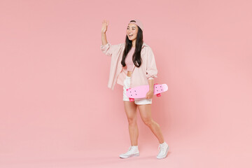 Full length portrait of funny young asian girl in casual clothes, cap isolated on pastel pink background. People lifestyle concept. Hold skateboard, waving and greeting with hand as notices someone.