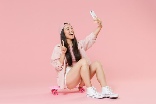 Full length portrait of cheerful young asian woman girl in cap isolated on pastel pink background. People lifestyle concept. Sit on skateboard, doing selfie shot on mobile phone, showing victory sign.