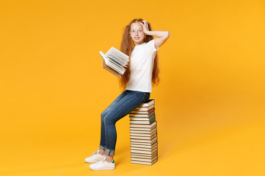Full length young redhead girl 12-13 years old in white t-shirt jeans sit on big stack school textbook notebook read book isolated on yellow background children studio portrait Kids education concept