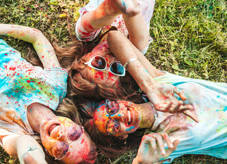 Three happy beautiful girls making party at Holi colors festival in summer time.Young  smiling women friends having fun after music event at sunset.Models lying on the grass in sunglasses