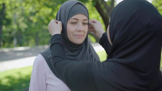 Unrecognizable woman adjusting hijab of smiling young beautiful lady outdoors. Portrait of confident Muslim female friends enjoying sunny summer day outdoors. Traditional culture concept.
