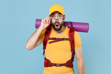 Shocked traveler young man in yellow casual t-shirt cap glasses with backpack isolated on blue background. Tourist traveling on weekend getaway. Tourism discovering hiking concept. Keeping mouth open.