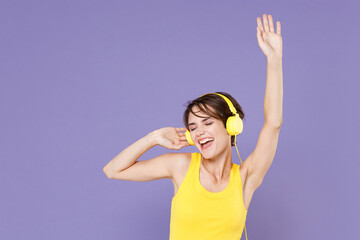 Cheerful young brunette woman girl wearing yellow casual tank top posing isolated on pastel violet background studio. People lifestyle concept. Listening music with headphones dancing rising hands.