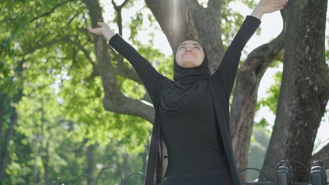Portrait of happy young Muslim woman in black traditional clothes and hijab enjoying sunny summer day in park. Smiling charming female immigrant resting outdoors. Freedom and lifestyle concept.