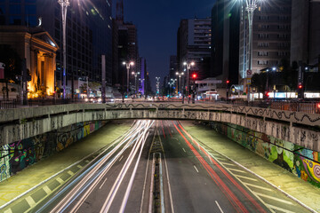Imagem em longa exposição noturna da Avenida Paulista com rastro das luzes dos veículos 
