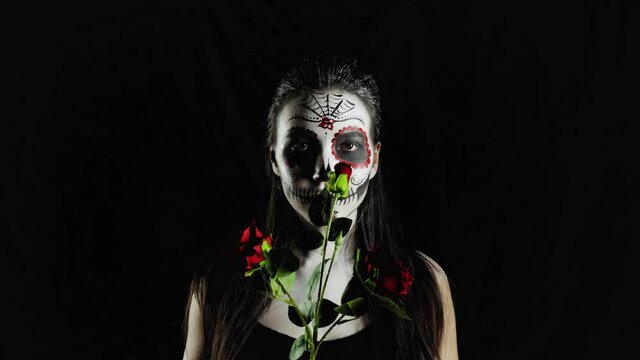 Halloween mask Calavera Catrina. Girl with a rose in her hands. Mexican day of the dead. Portrait of a young woman with a terrible multi-colored makeup for Halloween on a dark background. 4K. 