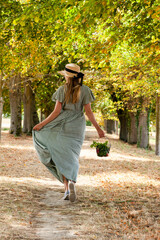 Beautiful happy young girl in a straw hat and long dress running in the forest with a basket in her hand. Summer or early autumn. Bright warm fall colors. Back view.