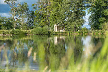 Fototapeta na wymiar A view with blurred grass in the foreground across a beautiful lake with reflections of trees towards an old abandoned structure in the middle of a rural countryside scene
