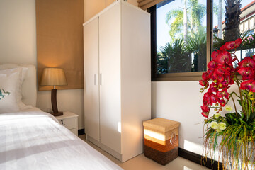 Interior design of house home, villa, condo, apartment feature white wardrobe, bedside table, lamp, basket, window glass and artificial flower in bedroom