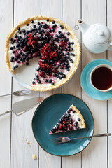 Blueberry open pie on blue plate with cup of black tea and white tea pot on bright painted wood background