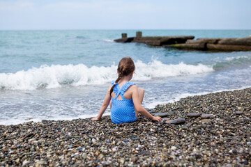 View from behind of toddler 5-6 year old girl in swimsuit sitting on the beach and playing with pebbles on sunny day summer family vacation with kids. children having fun on the sea