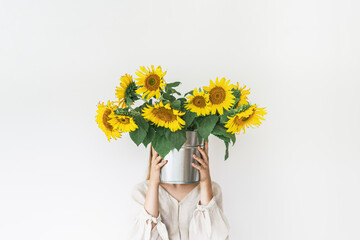 Beautiful young woman in linen dress holds sunflowers bouquet in a tin bucket on white background.
