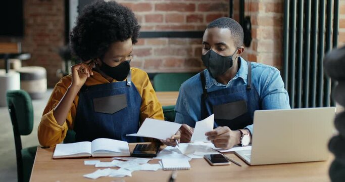 African American couple in masks doing business calculations of restaurant and checking bills. Calculating spends and damages. Reopen after lockdown. Counting at laptop computer Small entrepreneurship