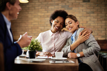 Happy female colleagues embracing with eyes closed while having coffee break in a cafe.