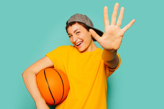 Portrait of cute girl in sporty outfit over blue background holding a basketball ball.