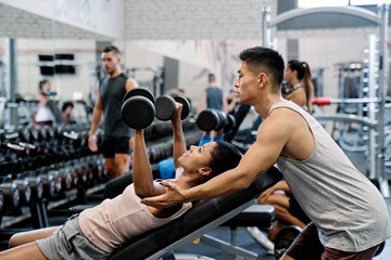 Fit dark skinned woman in sportswear doing a dumbbell bench press lying on an inclined bench with a asian male instructor partner in the gym.