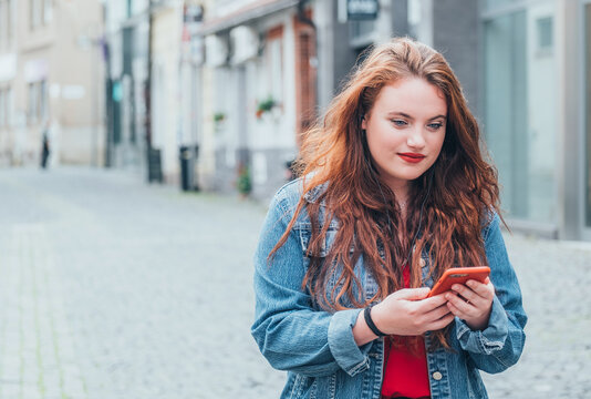 Portrait of red curled long hair caucasian teen girl walking on the street and browsing the internet using the modern smartphone. Modern people with technology devices concept image.