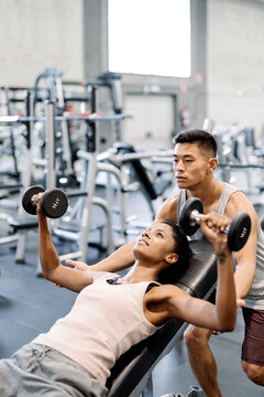 Fit dark skinned woman in sportswear doing a dumbbell bench press lying on an inclined bench with a asian male instructor partner in the gym.