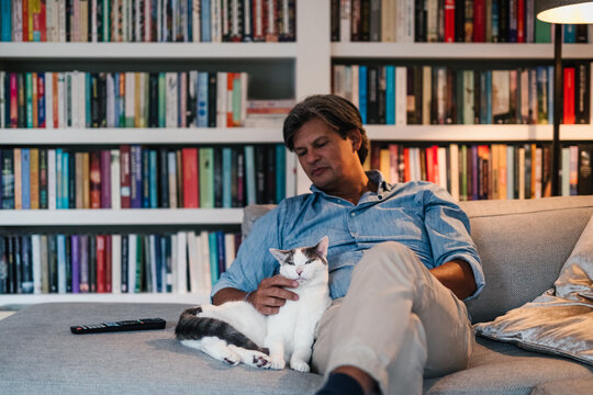 Portrait of a man sitting on the couch in front of a big bookcase, petting his cat